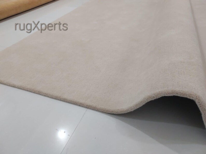 May include: A close-up of a beige rug with a soft, textured surface. The rug is partially rolled, revealing its thickness and construction. The text "rugXperts" is visible in the upper left corner.