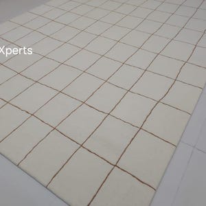 May include: White rug with a grid pattern of brown lines. The rug is made of wool and has a soft, luxurious feel.