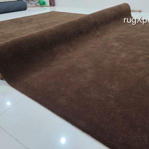 May include: A rolled-up, brown rug with a soft, textured surface. The rug is a solid color and appears to be made of a plush material. The brand name "rugXperts" is visible in the upper right corner. The rug is on a white tiled floor.