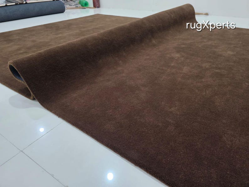May include: A rolled-up, brown rug with a soft, textured surface. The rug is a solid color and appears to be made of a plush material. The brand name "rugXperts" is visible in the upper right corner. The rug is on a white tiled floor.