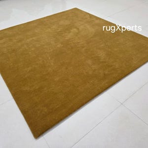 May include: A mustard-coloured rectangular rug with a soft, textured surface. The rug is placed on a white tiled floor. The text "rugXperts" is visible in the upper right corner.