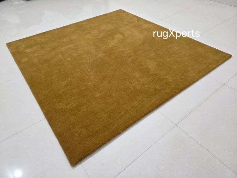 May include: A mustard-coloured rectangular rug with a soft, textured surface. The rug is placed on a white tiled floor. The text "rugXperts" is visible in the upper right corner.