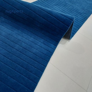 Op de afbeelding: Een opgerolde, lange, blauwe loper met een geribbelde textuur. De loper heeft een diepe blauwe tint en lijkt gemaakt te zijn van een zacht materiaal. De loper ligt op een witte tegelvloer. De merknaam "rugXperts" is zichtbaar.