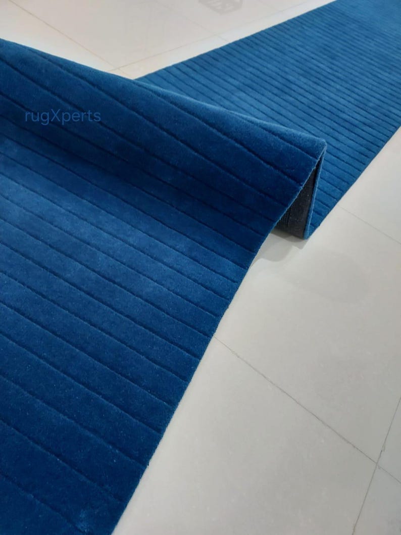 Op de afbeelding: Een opgerolde, lange, blauwe loper met een geribbelde textuur. De loper heeft een diepe blauwe tint en lijkt gemaakt te zijn van een zacht materiaal. De loper ligt op een witte tegelvloer. De merknaam "rugXperts" is zichtbaar.