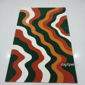May include: A rectangular rug with a wavy pattern in shades of green, white, orange, and rust. The rug's design features flowing, organic lines that create a visually dynamic effect. The rug is likely made of a soft material, suitable for a living room or bedroom.