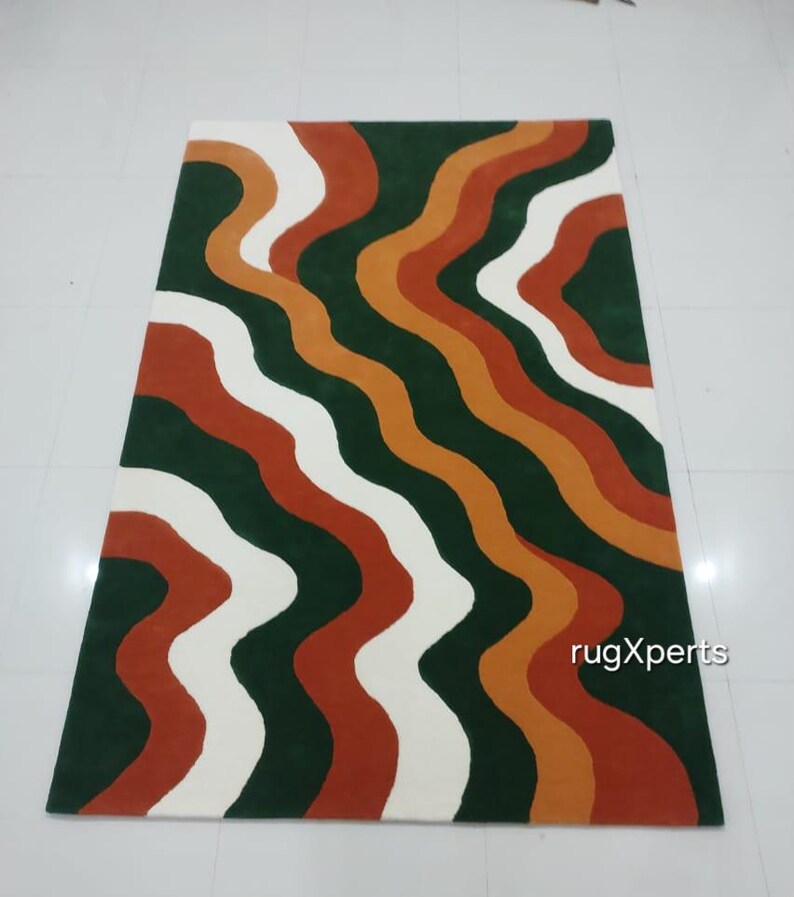 May include: A rectangular rug with a wavy pattern in shades of green, white, orange, and rust. The rug's design features flowing, organic lines that create a visually dynamic effect. The rug is likely made of a soft material, suitable for a living room or bedroom.