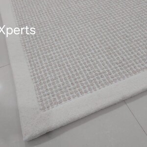 May include: Close-up of a white woven rug with a white border. The rug has a subtle texture and a slightly off-white color.