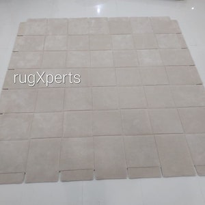 Pu&ograve; includere: Un tappeto beige fatto di quadrati singoli, con il testo "rugXperts" al centro.