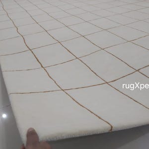 May include: White rug with a grid pattern of brown lines. The rug is folded in half and the text "rugXperts" is visible in the lower right corner.