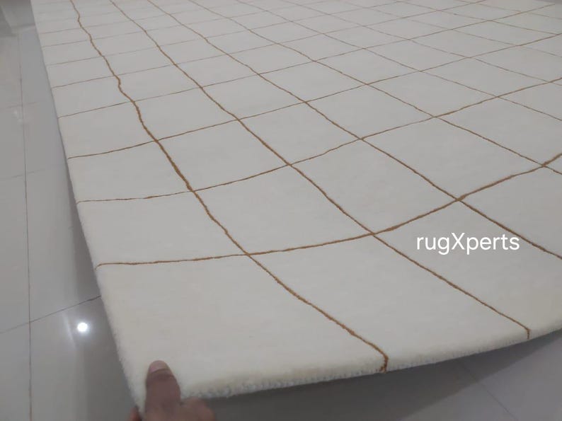 May include: White rug with a grid pattern of brown lines. The rug is folded in half and the text "rugXperts" is visible in the lower right corner.
