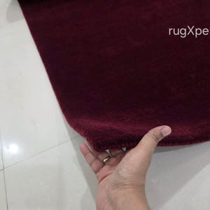 May include: A close-up of a burgundy rug with a soft, textured surface. The rug is being held by a hand. The text "rugXperts" is visible in the upper right corner.