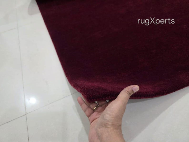 May include: A close-up of a burgundy rug with a soft, textured surface. The rug is being held by a hand. The text "rugXperts" is visible in the upper right corner.