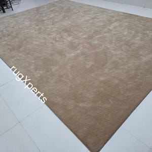 May include: A large, rectangular, beige rug with a soft, textured surface. The rug is placed on a tiled floor. The words "rugXperts" are printed on the rug.