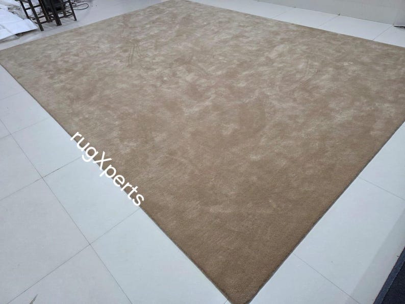May include: A large, rectangular, beige rug with a soft, textured surface. The rug is placed on a tiled floor. The words "rugXperts" are printed on the rug.