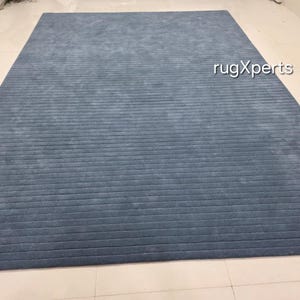 Op de afbeelding: Een groot, rechthoekig blauw tapijt met een gestructureerd, geribbeld ontwerp. Het tapijt heeft een effen kleur, met een licht vervaagde uitstraling. Het tapijt ligt op een betegelde vloer. De tekst "rugXperts" is zichtbaar op het tapijt.