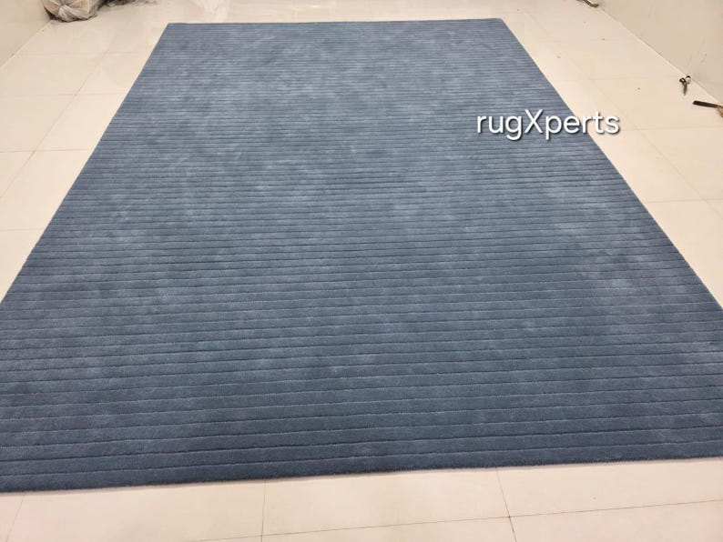 Op de afbeelding: Een groot, rechthoekig blauw tapijt met een gestructureerd, geribbeld ontwerp. Het tapijt heeft een effen kleur, met een licht vervaagde uitstraling. Het tapijt ligt op een betegelde vloer. De tekst "rugXperts" is zichtbaar op het tapijt.