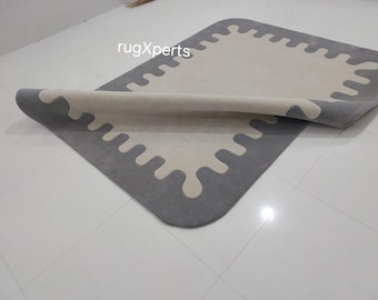 Puede incluir: Una alfombra gris y blanca con un diseño abstracto único. La alfombra tiene un borde gris con un patrón blanco y ondulado en el interior.