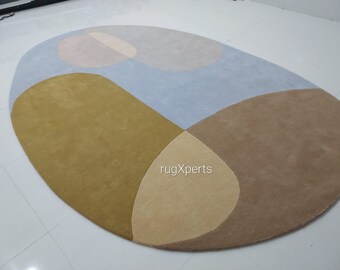 May include: An oval-shaped area rug with a modern design. The rug features a color-blocked pattern with sections of light blue, tan, brown, and mustard yellow. The rug is made of a soft material and is suitable for a living room or bedroom.