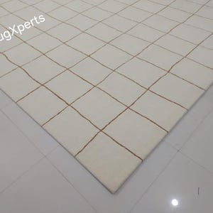 May include: A white rug with a grid pattern of thin brown lines. The rug is shown from a corner view.