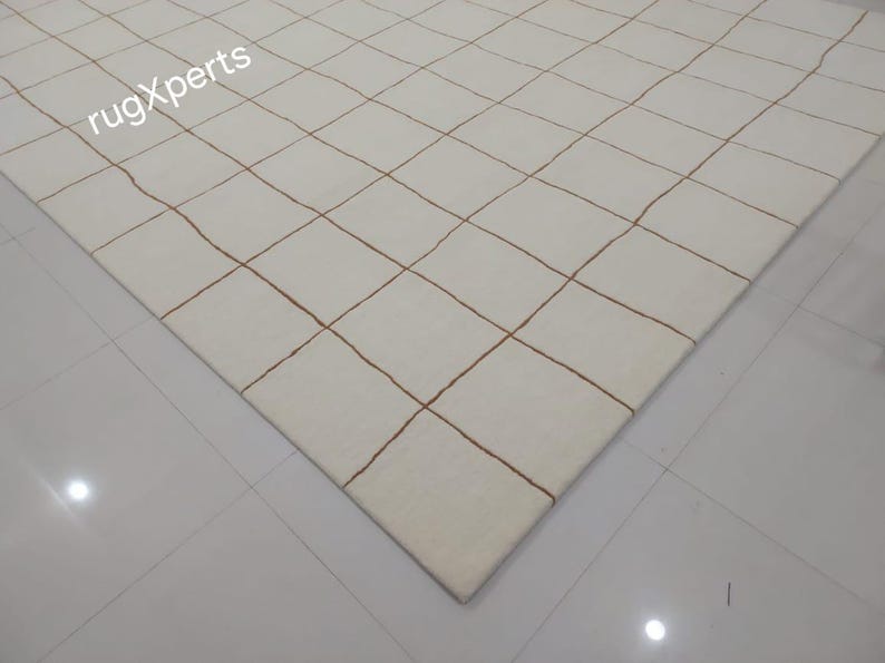 May include: A white rug with a grid pattern of thin brown lines. The rug is shown from a corner view.