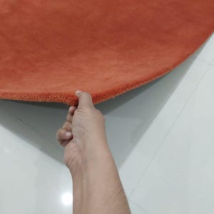 May include: A round, burnt orange rug with a soft, textured surface. The edge of the rug is held up, revealing the thickness and construction. The rug is placed on a white tiled floor.
