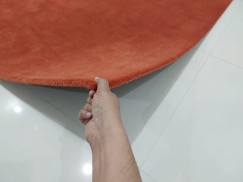 May include: A round, burnt orange rug with a soft, textured surface. The edge of the rug is held up, revealing the thickness and construction. The rug is placed on a white tiled floor.