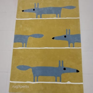 May include: A yellow rug with a repeating pattern of three blue stylized foxes. The rug has a white border and the text "rugXperts" is printed in the bottom left corner.