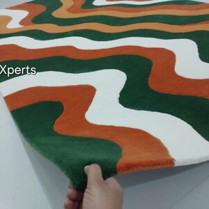 May include: A rectangular rug with a wavy pattern in green, white, and orange. The rug's design features flowing, organic shapes that create a visually appealing and modern aesthetic. The rug is made of a soft, plush material.