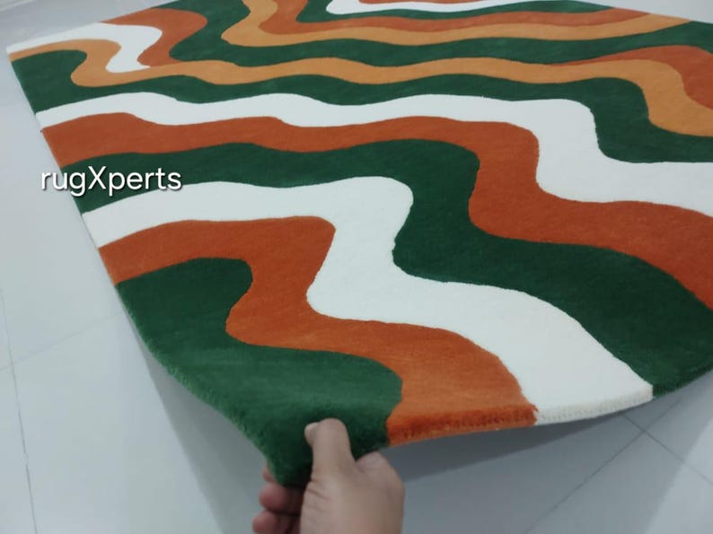 May include: A rectangular rug with a wavy pattern in green, white, and orange. The rug's design features flowing, organic shapes that create a visually appealing and modern aesthetic. The rug is made of a soft, plush material.