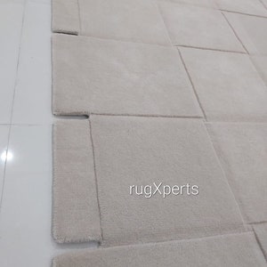 Pu&ograve; includere: Un primo piano di un tappeto beige chiaro con un motivo geometrico unico. Il tappeto &egrave; realizzato in un materiale morbido e soffice e ha una lucentezza sottile. Il tappeto &egrave; tagliato in quadrati e i bordi sono cuciti insieme per creare un design unico. Il tappeto &egrave; perfetto per aggiungere un tocco di stile moderno a qualsiasi stanza.
