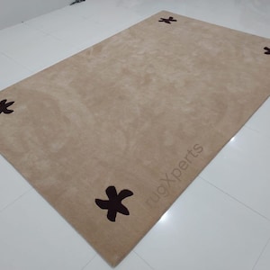 May include: A rectangular beige rug with four dark brown star-shaped designs in each corner. The rug has a soft, plush texture and is placed on a white tiled floor. The text "rugXperts" is visible on the rug.
