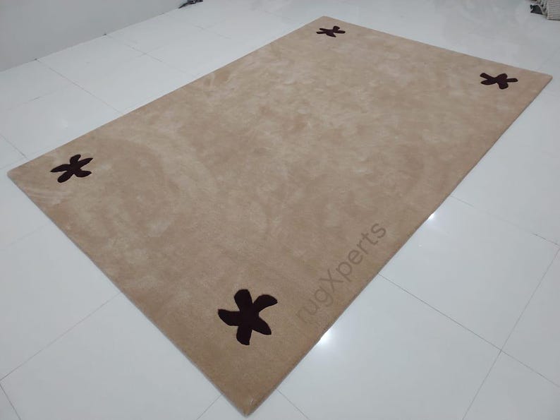 May include: A rectangular beige rug with four dark brown star-shaped designs in each corner. The rug has a soft, plush texture and is placed on a white tiled floor. The text "rugXperts" is visible on the rug.