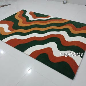 May include: A rectangular rug with a wavy pattern in shades of green, orange, white, and rust. The rug's design features horizontal, flowing lines, creating a modern and artistic look. The rug is placed on a white tiled floor.