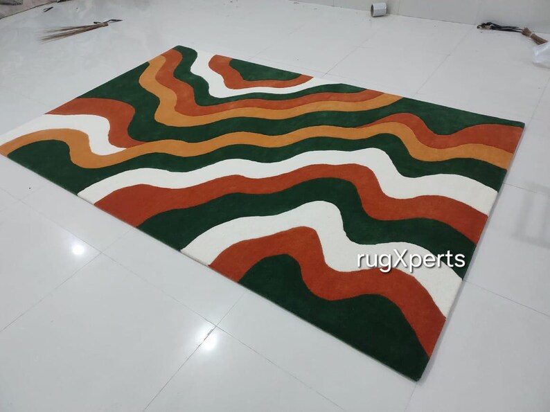 May include: A rectangular rug with a wavy pattern in shades of green, orange, white, and rust. The rug's design features horizontal, flowing lines, creating a modern and artistic look. The rug is placed on a white tiled floor.