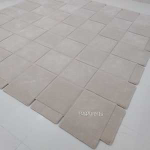 Pu&ograve; includere: Un grande tappeto modulare di colore avorio, composto da quadrati singoli che possono essere disposti in diversi motivi. Il tappeto &egrave; realizzato in un materiale morbido e soffice e presenta una texture sottile. Il tappeto &egrave; mostrato su un pavimento bianco. Il testo "rugXperts" &egrave; visibile nell'angolo in basso a destra dell'immagine.