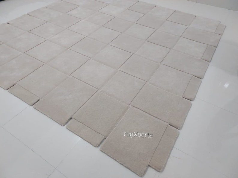 Pu&ograve; includere: Un grande tappeto modulare di colore avorio, composto da quadrati singoli che possono essere disposti in diversi motivi. Il tappeto &egrave; realizzato in un materiale morbido e soffice e presenta una texture sottile. Il tappeto &egrave; mostrato su un pavimento bianco. Il testo "rugXperts" &egrave; visibile nell'angolo in basso a destra dell'immagine.