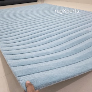 May include: A light blue, rectangular rug with a ribbed design. The rug's surface has a soft, plush texture, and the pattern consists of parallel, curved lines. The rug is displayed on a light-colored floor.