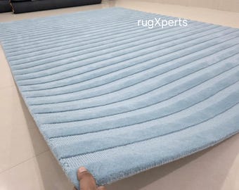 May include: A light blue, rectangular rug with a ribbed design. The rug's surface has a soft, plush texture, and the pattern consists of parallel, curved lines. The rug is displayed on a light-colored floor.