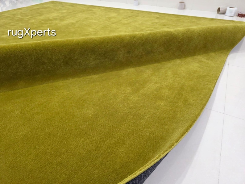 May include: A large, rectangular, olive-green rug with a soft, plush texture. The rug is partially rolled up, revealing a dark grey backing. The text "rugXperts" is visible in the upper left corner. The rug is on a white surface.
