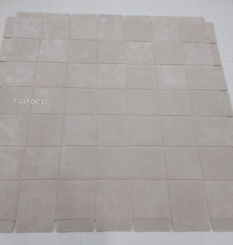 Pu&ograve; includere: Un grande tappeto quadrato di colore bianco sporco, composto da quadrati singoli cuciti insieme. Il tappeto &egrave; realizzato in un materiale morbido e soffice.