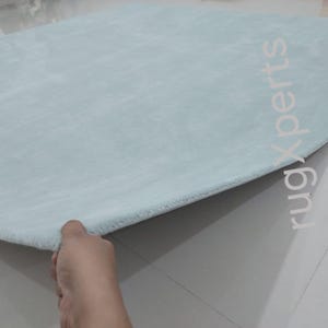 May include: A light blue, oval-shaped rug with a soft, plush texture. The rug is being held up at the edge, revealing its thickness and construction. The background is a light-colored tiled floor.
