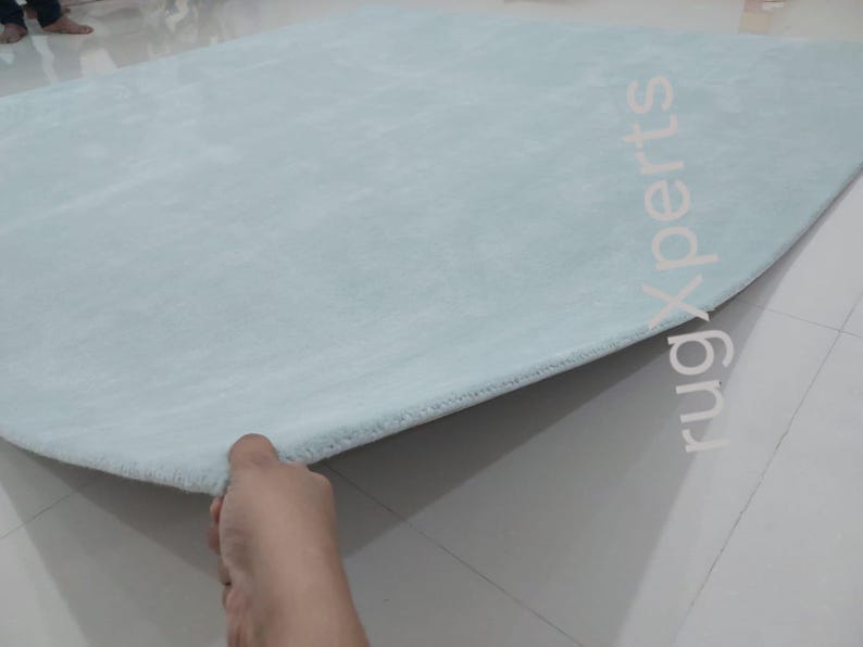 May include: A light blue, oval-shaped rug with a soft, plush texture. The rug is being held up at the edge, revealing its thickness and construction. The background is a light-colored tiled floor.