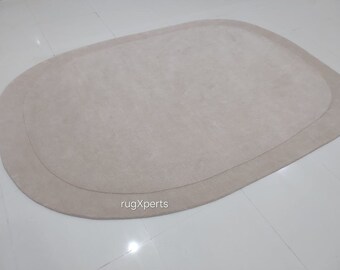 Könnte beinhalten: Ein hellbeiger ovaler Teppich mit einem etwas dunkleren beigefarbenen Rand. Der Teppich besteht aus einem weichen, plüschigen Material und hat einen dezenten Glanz. Der Teppich ist auf einem weißen Fliesenboden fotografiert. Der Text "rugXperts" ist in der unteren rechten Ecke des Bildes sichtbar.