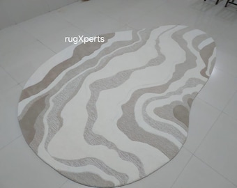 May include: A large, abstract, off-white rug with a wavy, organic pattern in shades of beige. The rug is shaped like a teardrop and has a soft, plush texture.