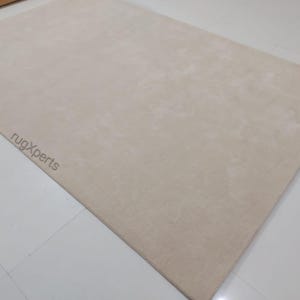 May include: A large, rectangular beige rug. The rug has a soft, plush texture and is laid out on a white tiled floor. The words "rugXperts" are subtly printed in the lower left corner.
