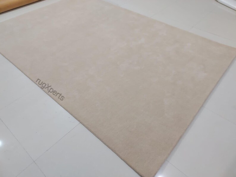 May include: A large, rectangular beige rug. The rug has a soft, plush texture and is laid out on a white tiled floor. The words "rugXperts" are subtly printed in the lower left corner.