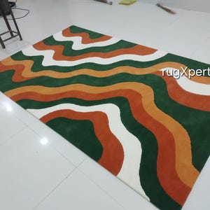 May include: A rectangular rug with a wavy pattern in shades of green, orange, white, and rust. The rug has a modern design with the text "rugXperts" in the upper right corner. The rug is placed on a white tiled floor.