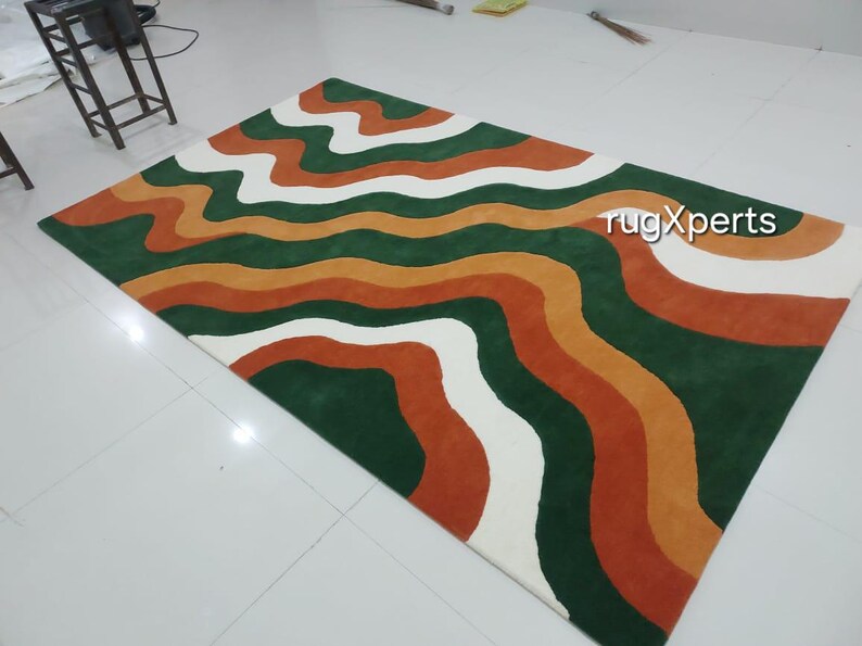 May include: A rectangular rug with a wavy pattern in shades of green, orange, white, and rust. The rug has a modern design with the text "rugXperts" in the upper right corner. The rug is placed on a white tiled floor.