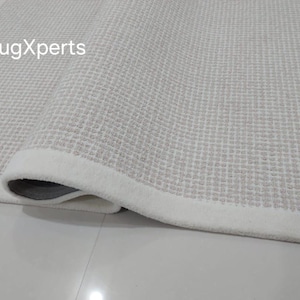 May include: Close-up of a white rug with a subtle textured pattern. The rug has a white border and is rolled up, showing the underside.