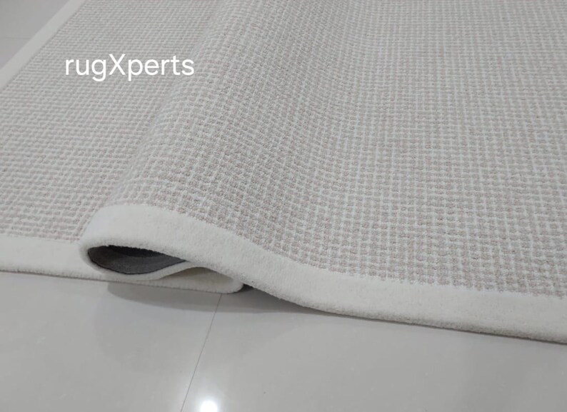 May include: Close-up of a white rug with a subtle textured pattern. The rug has a white border and is rolled up, showing the underside.